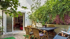 Un patio con mesa y sillas, rodeado de follaje y una pared con plantas colgantes.