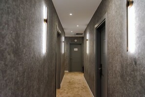 Um corredor com piso carpeteado, paredes com painéis texturizados e portas com placas iluminadas.