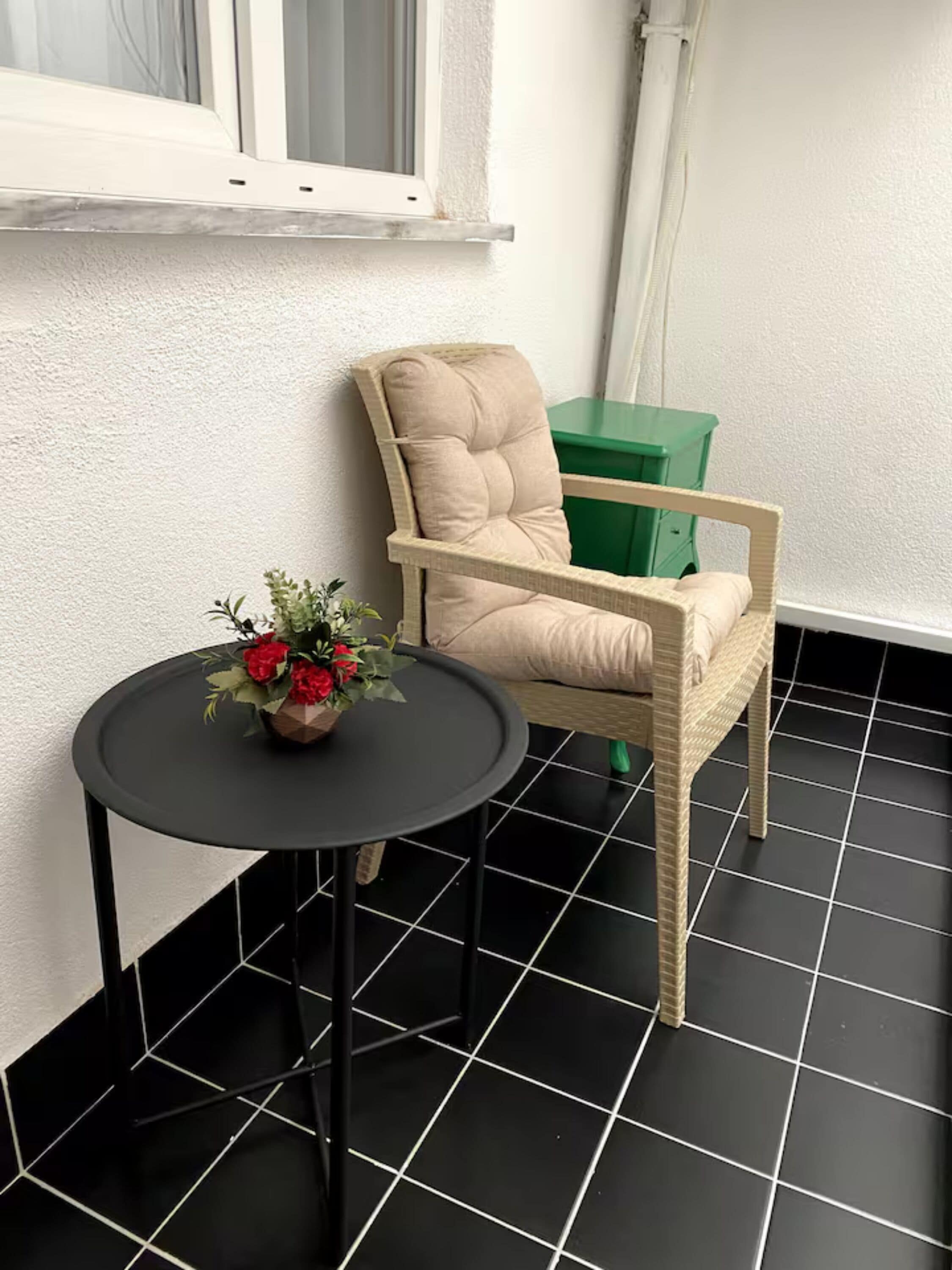 A room with a black tiled floor, a beige armchair, a small round table with a flower vase, and a green storage unit.