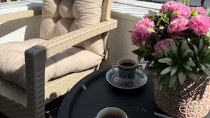 Una silla de mimbre con cojines, una mesa con una taza de café y una planta en maceta, y una barandilla de balcón.
