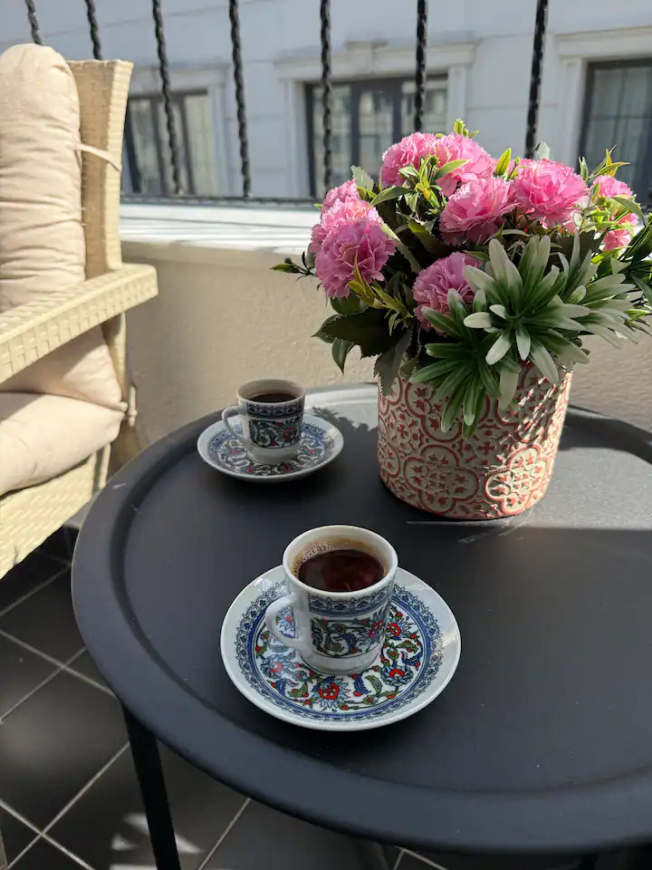 A small round table with a cup of tea and a matching saucer, a decorative vase with pink flowers, and a wicker chair.