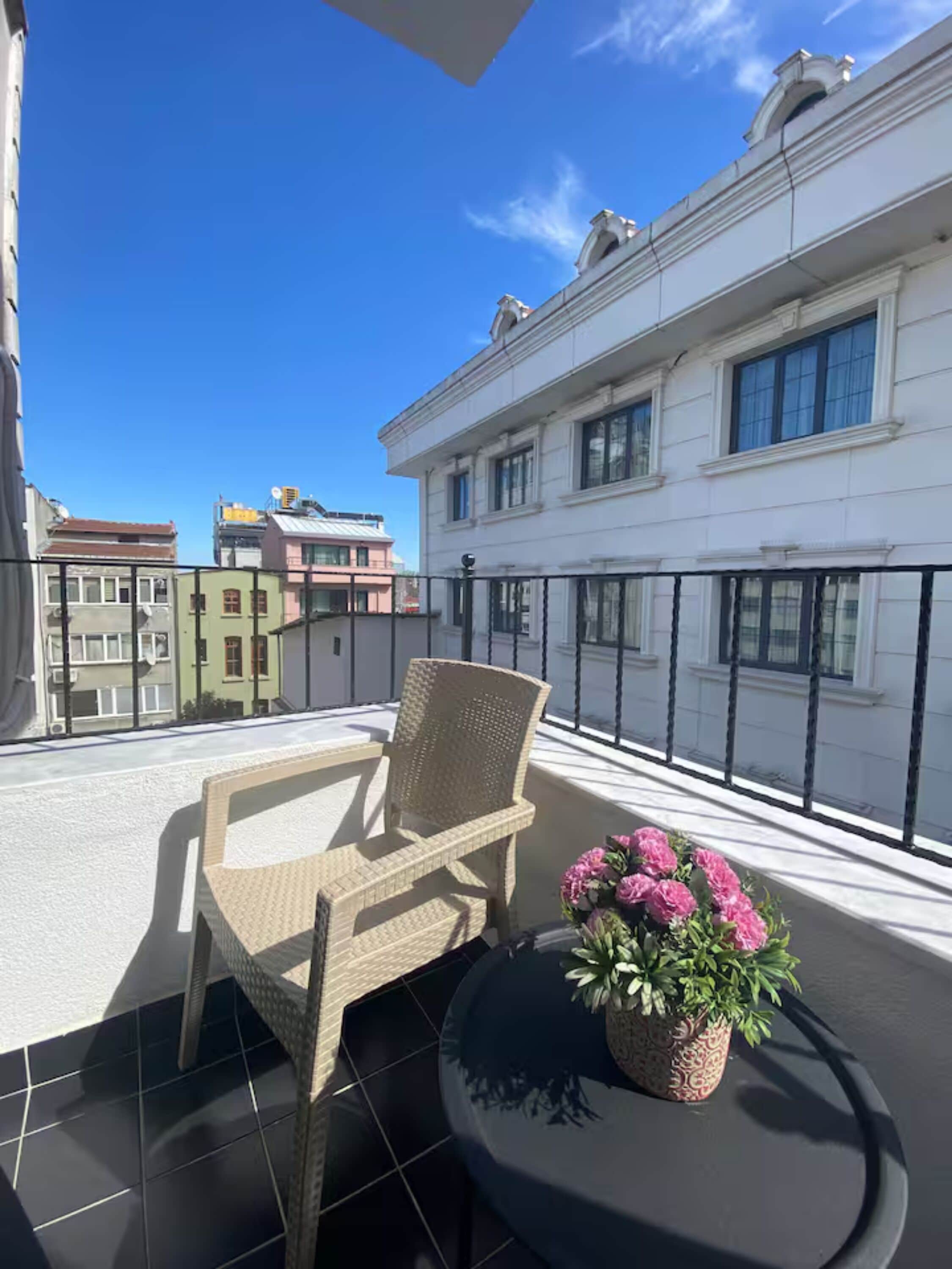 Un balcon avec une chaise en osier et une table, un pot de fleurs et une vue sur les immebles.