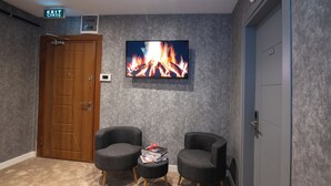 Una habitación con puerta de madera, televisor de pantalla plana mostrando fuego, dos sillones, una mesita con libros y suelo alfombrado.