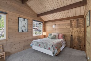 Ein Schlafzimmer mit Holzbalkendecke, einem Bett mit floralem Bettzeug, einem Nachttisch und einem Fenster mit Blick auf Bäume.