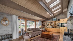 A spacious living room with a wooden ceiling, large windows, a fireplace, and a library. - Columbia Gorge - Mosier Bluff | River Views & Pool (Mosier)
