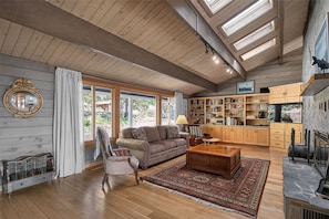 Ein geräumiges Wohnzimmer mit Holzboden, großen Fenstern, einem Kamin und einer Bibliothek.