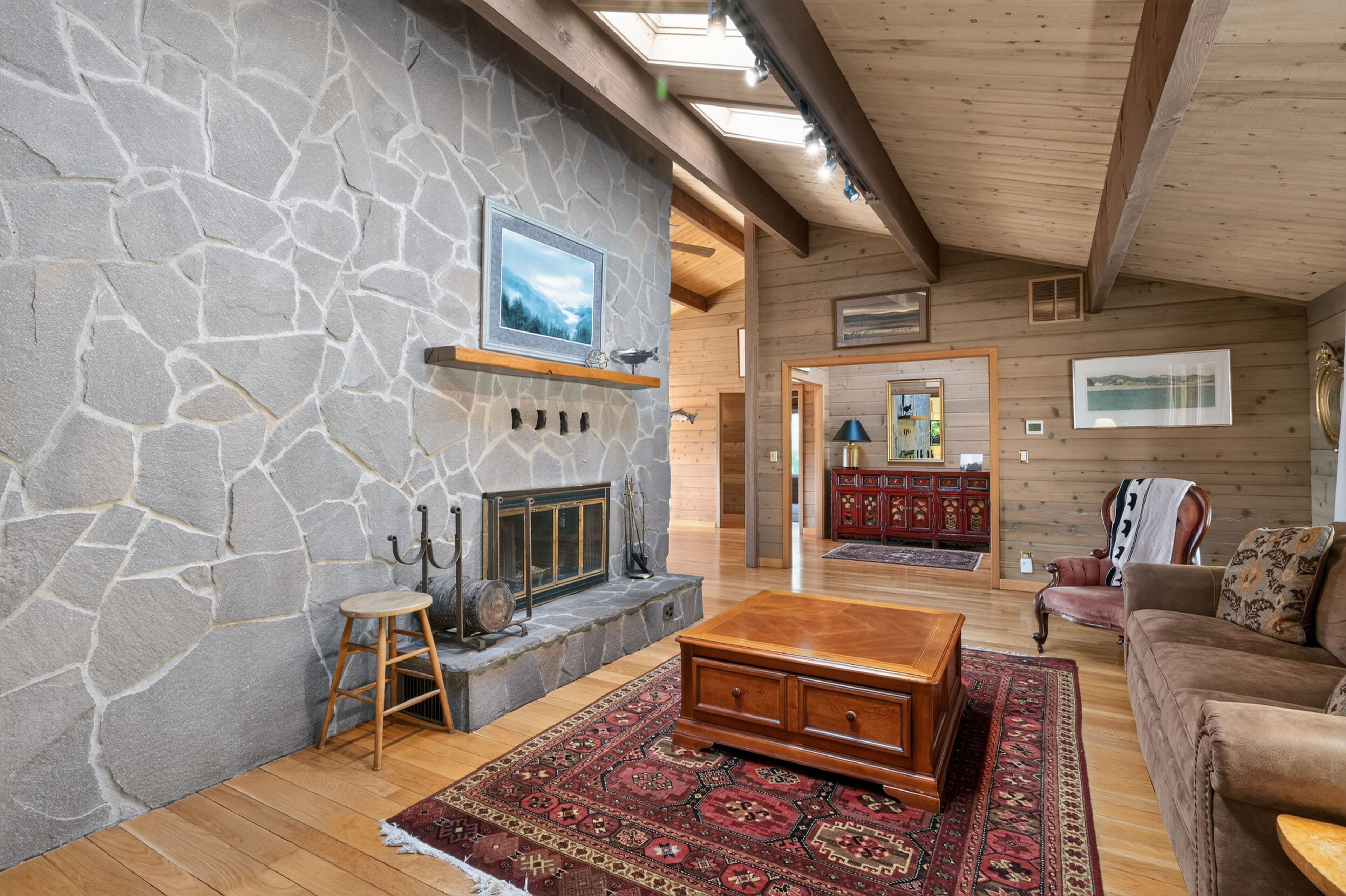 Une cheminée en pierre avec un téléviseur fixé au mur, un banc en bois, un tapis à motifs et un meuble en bois dans une pièce chaleureuse, dotée de poutres apparentes et d’un plafond en pente.