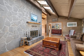 A stone fireplace with a mounted television, a wooden bench, a patterned rug, and a wooden cabinet in a cozy room with wooden beams and a sloped ceiling.