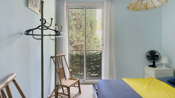 Ein Schlafzimmer mit einem Bett, einem Sessel, einem Kleiderständer, einem Fenster mit Vorhängen und einem Ventilator.