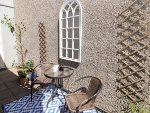 An outdoor patio with a round table, two wicker chairs, a potted plant, and a window with a lattice design.