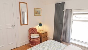 Un dormitorio con cama, una silla, una mesita de noche, una lámpara, una ventana con cortinas y una puerta.