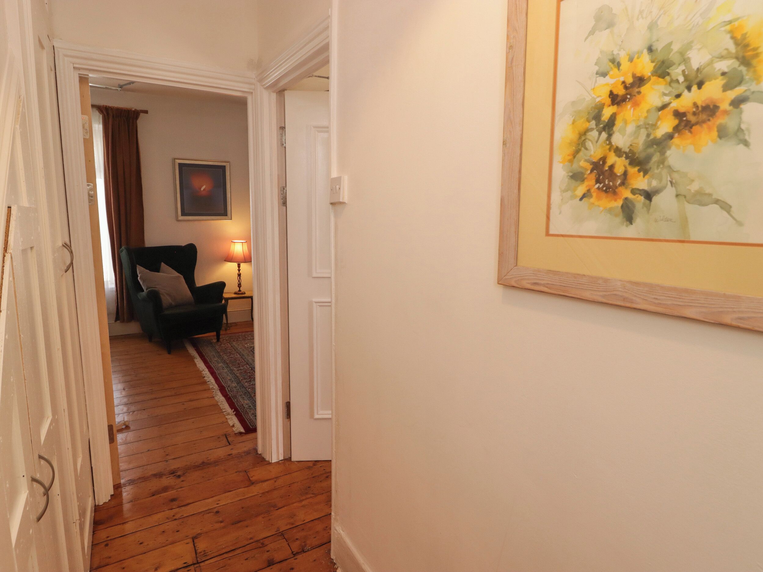 A hallway with wooden flooring leading to a room with a green armchair and a lamp.