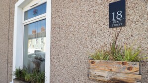 Fachada de edificio con una ventana, una maceta de madera con plantas y una señal de calle que indica '18 Kent Street'.