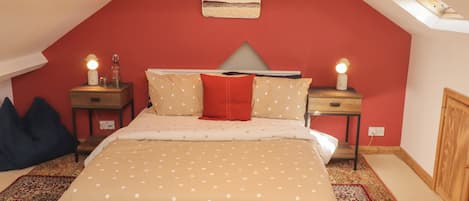 Un dormitorio con techo inclinado, una cama con ropa de cama de lunares, dos lámparas de noche, un cojín rojo y una mesita con una lámpara.