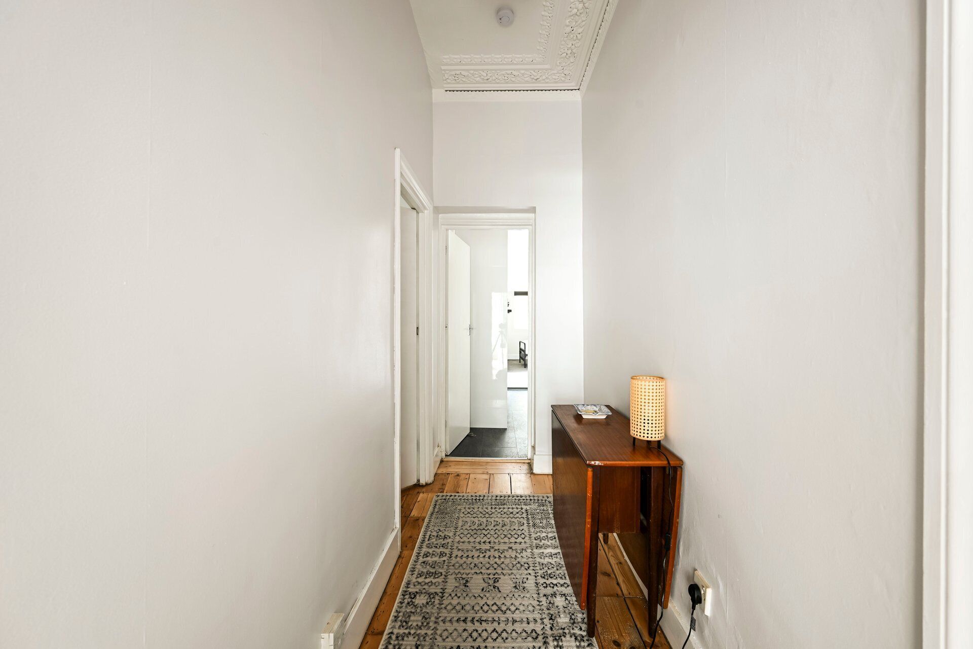 A hallway with a patterned rug, a wooden side table with a woven lamp, and a door leading to another room.