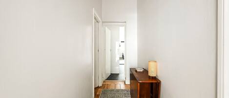 A hallway with a patterned rug, a wooden side table with a woven lamp, and a door leading to another room.