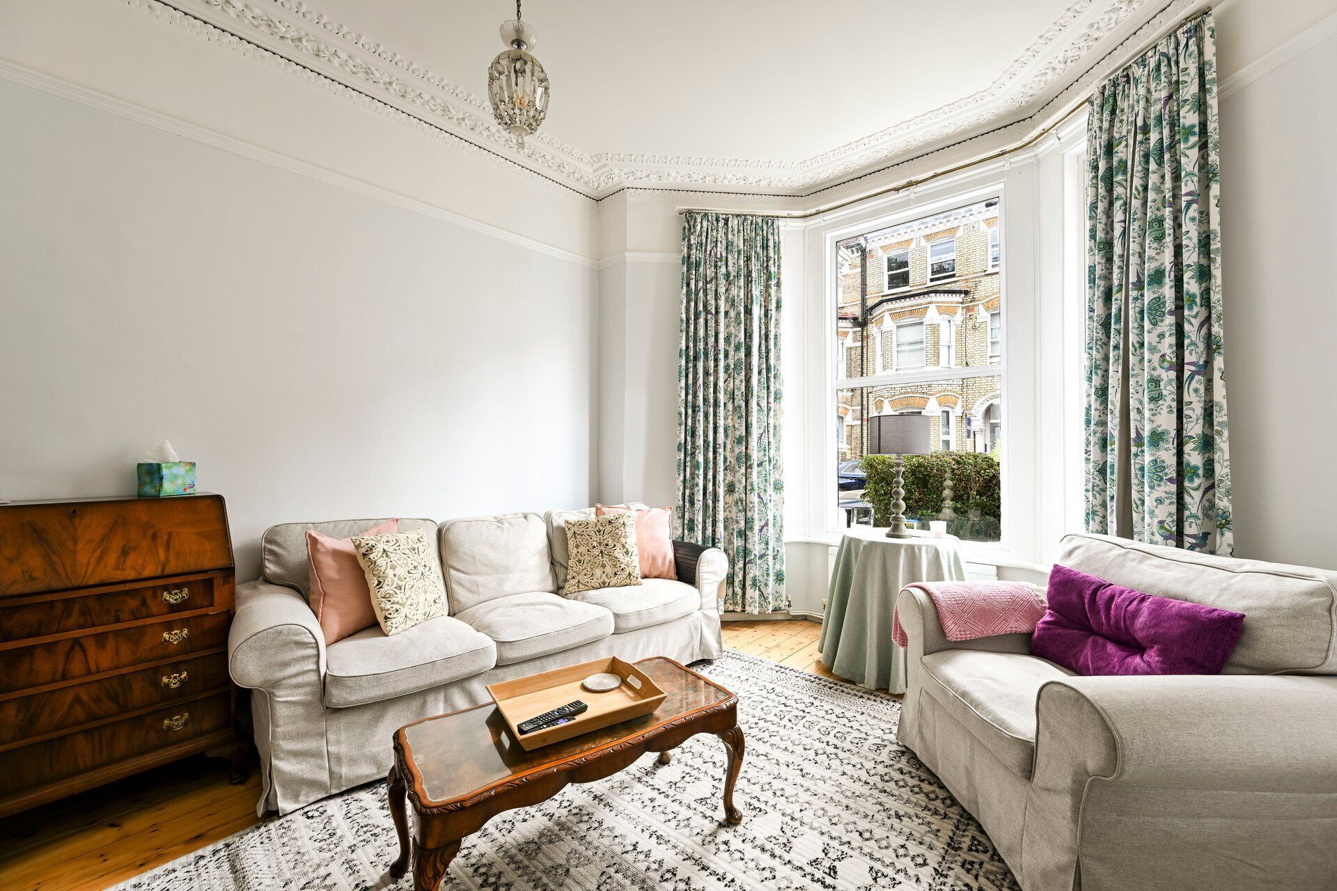 A living room with two sofas, a wooden coffee table, and a view of a building through a window with patterned curtains.