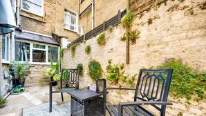 An outdoor seating area with two chairs and a table, surrounded by potted plants and a textured wall. - GuestReady - Stylish 1bed w Backyard in Clapham (London)
