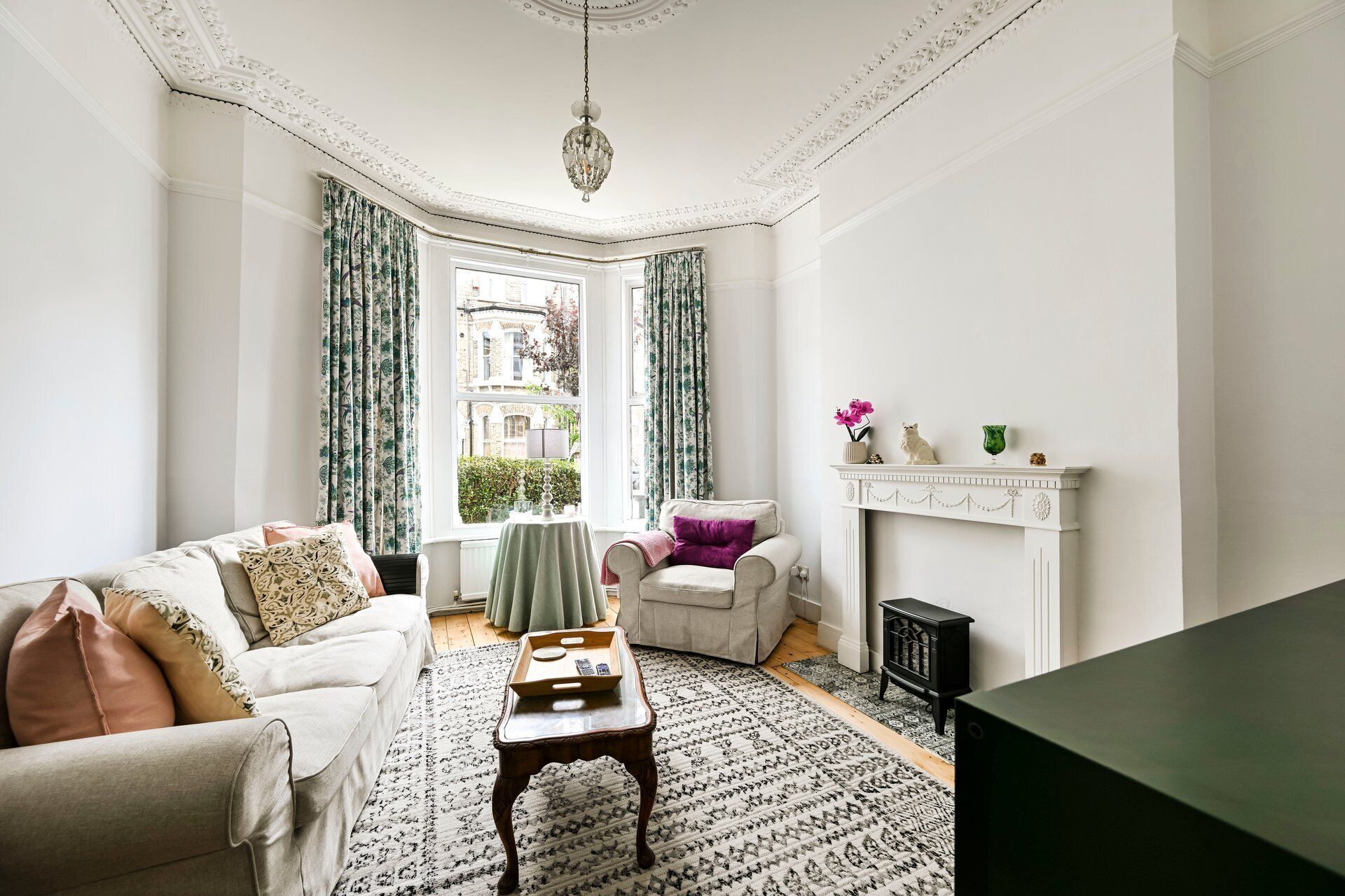Ein Zimmer mit einem weißen Sofa, einem gemusterten Teppich, einem kleinen Tischchen mit Tablett, einem Sessel mit violettem Kissen, einem Kamin mit Dekoration und Blick auf ein Gebäude durch ein Fenster mit Vorhängen.