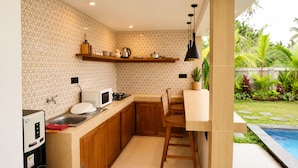 A compact kitchen with a microwave, stove, and wooden cabinets, adjacent to a dining area with a pool view. - Eka Wisnu 2BR Private Pool Villa by TVM (Ubud)