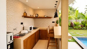 A compact kitchen with a microwave, stove, and wooden cabinets, adjacent to a dining area with a pool view. - Eka Wisnu 2BR Private Pool Villa by TVM (Ubud)