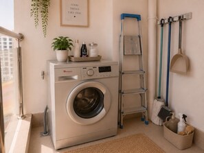 A compact laundry room with a front-loading washing machine, a metal drying rack, a broom, a dustpan, and a small plant on the counter. - Modern City Escape Near JKIA, Rooftop Pool, Gym & Free Parking (Nairobi)