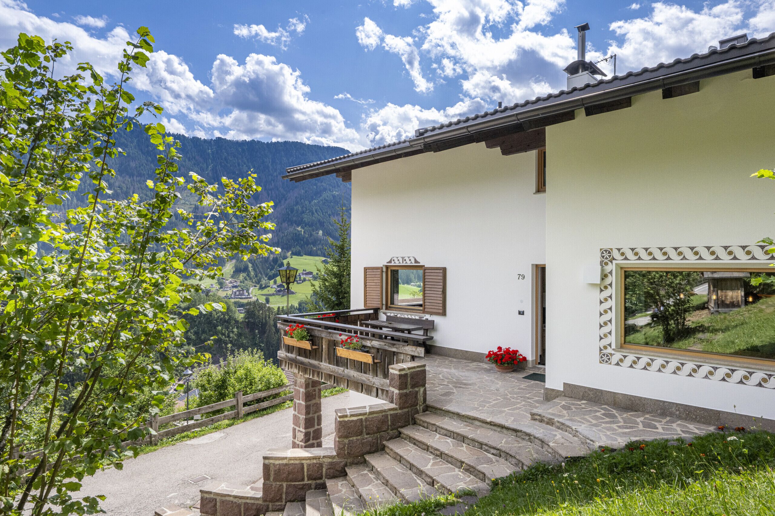 A modern building with a stone staircase leading to a balcony, a view of a mountain landscape, and a small garden area with flowers.