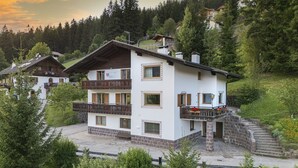 A multi-story chalet with a balcony, stone foundation, and a view of a forested hillside. - 1015A Valdussa Panoramic Apartment (Ortisei)