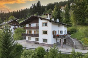 Ein mehrstöckiges Chalet mit Balkon, Steinuntergrund und Blick auf einen bewaldeten Hügel.