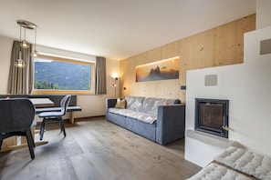 A modern living room with a sofa, a dining table with chairs, a fireplace, and a large window with a view of mountains.