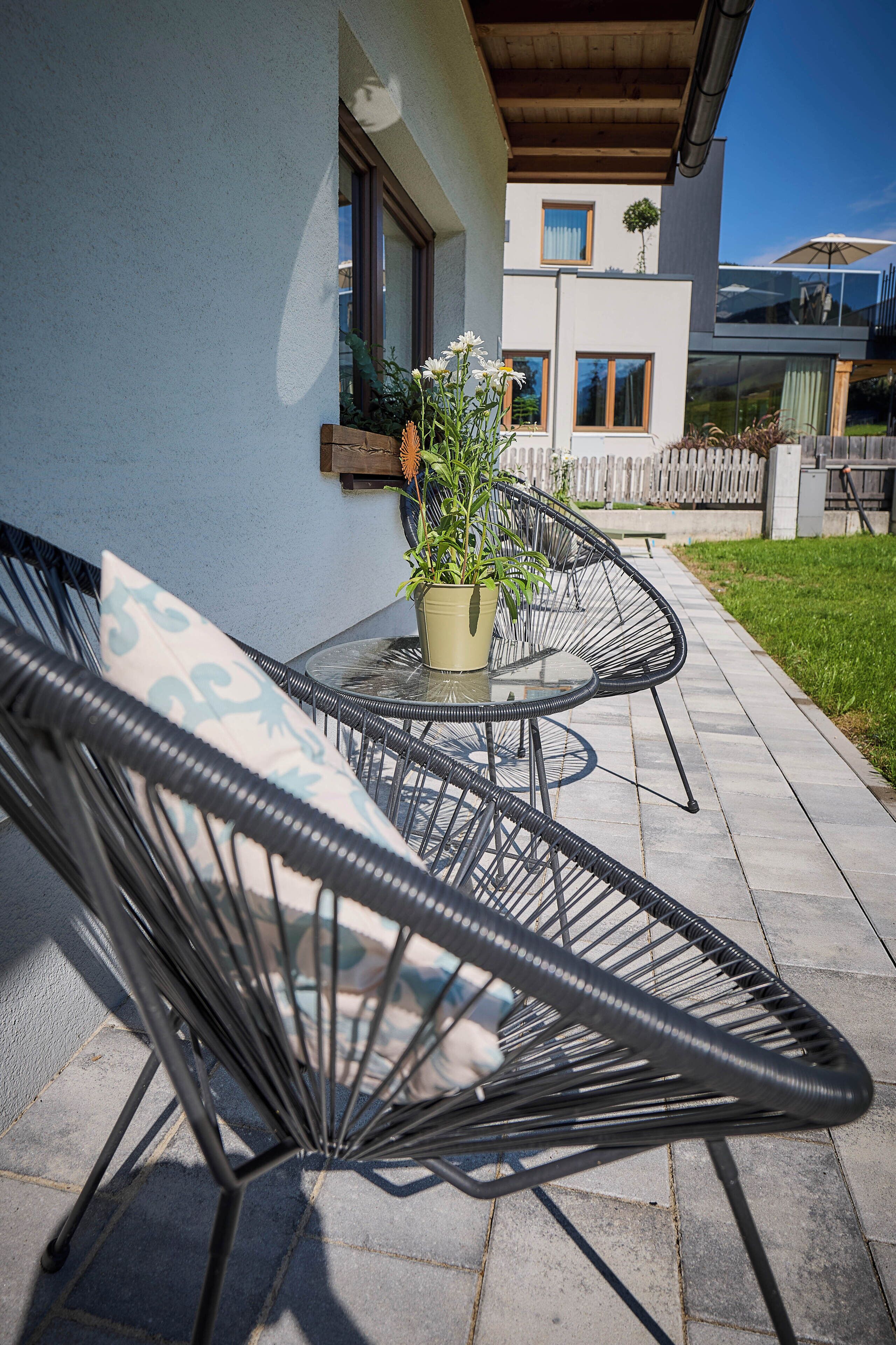 Een terras met twee zwarte stoelen met draadframe en een tafel, een potplant en een huis op de achtergrond.