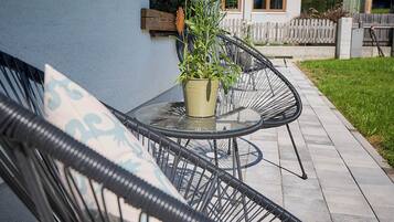Een terras met twee zwarte stoelen met draadframe en een tafel, een potplant en een huis op de achtergrond.