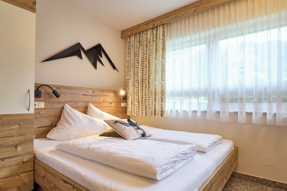 A hotel room with a wooden headboard, a bed with white linens, decorative pillows, a wall-mounted light fixture, and sheer curtains.