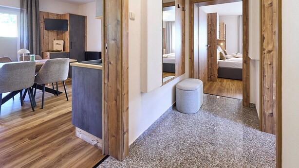 A hotel room with a wooden floor, a grey sofa, a small round ottoman, a bed with pillows, and a bathroom visible through an open door. - BERGLIEBE - Boutique Apartment Saalbach (Saalbach)