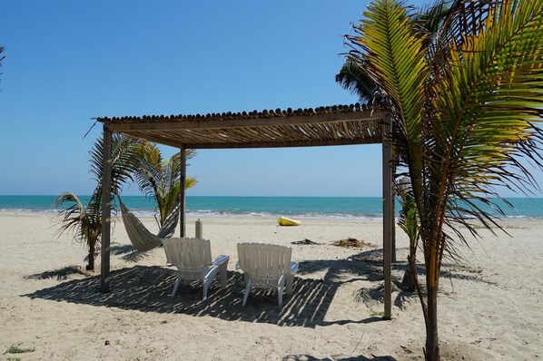 A beach scene with a thatched roof structure, two white chairs, and palm trees.