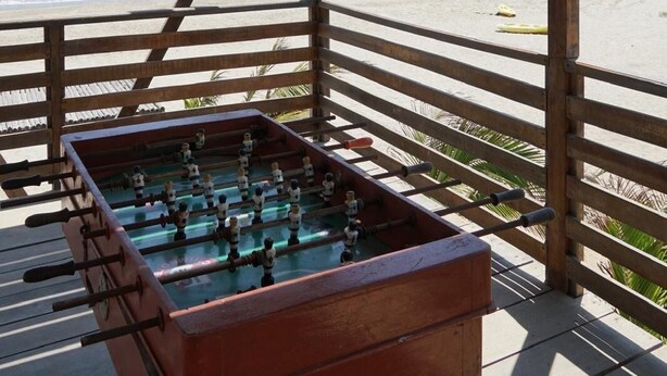 Zona de estar al aire libre con mesas de madera y una mesa de futbolín, con vista a una playa.