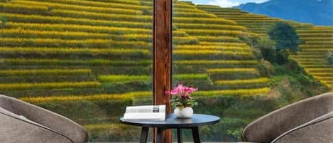 A room with a view of terraced fields, a wooden pole, a table with a book and flowers, and two chairs.
