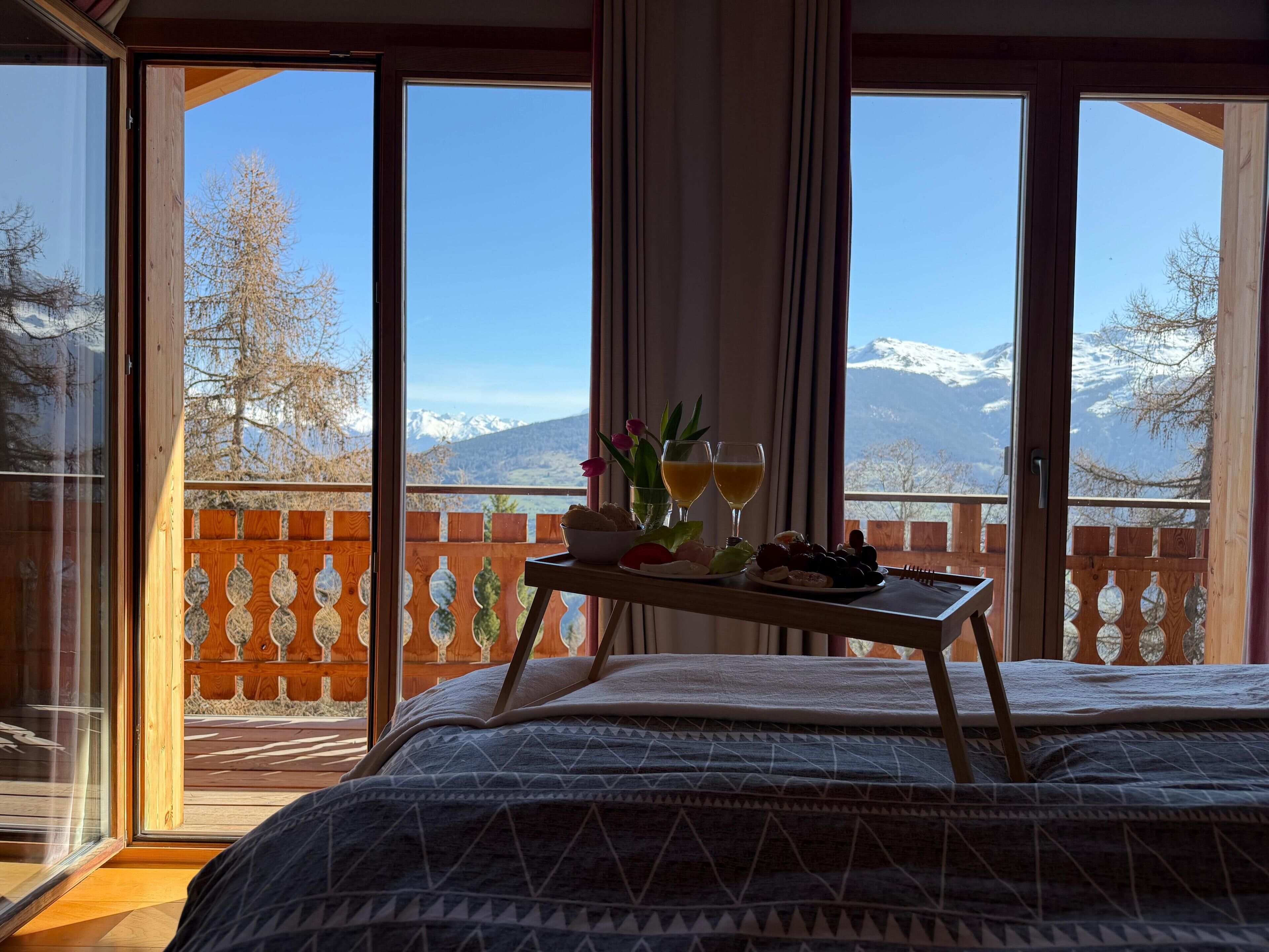 Une chambre avec un lit, une petite table d’appoint avec de la nourriture et des boissons, et une vue sur les montagnes par de grandes fenêtres.