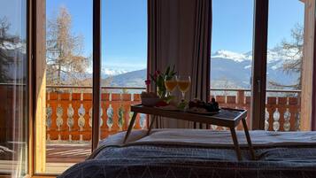 Une chambre avec un lit, une petite table d’appoint avec de la nourriture et des boissons, et une vue sur les montagnes par de grandes fenêtres.