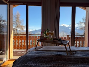 Une chambre avec un lit, une petite table d’appoint avec de la nourriture et des boissons, et une vue sur les montagnes par de grandes fenêtres.