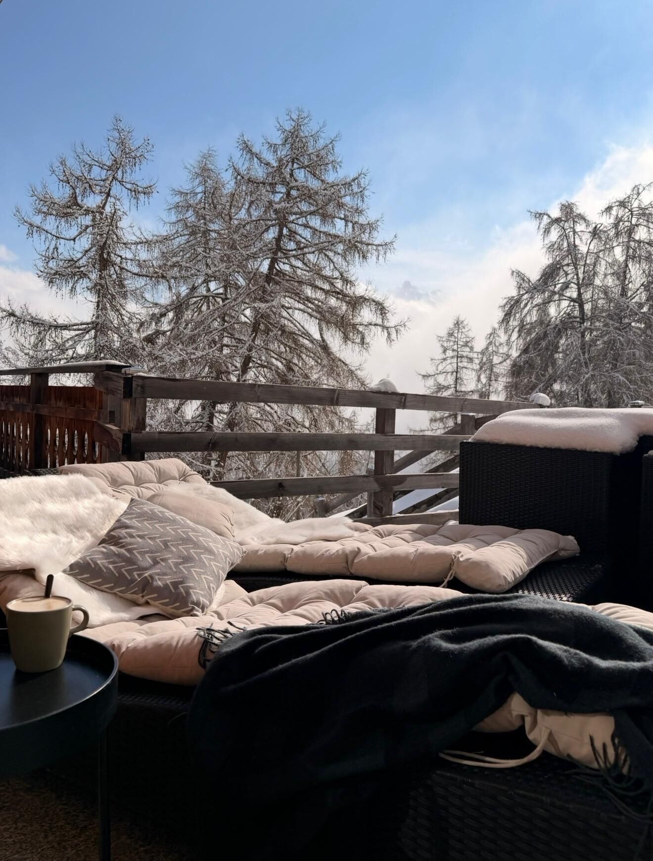 Un espace extérieur accueillant, aménagé avec un banc rembourré et une table, entouré d’une clôture en bois et d’arbres couverts de neige.