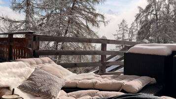 Un espace extérieur accueillant, aménagé avec un banc rembourré et une table, entouré d’une clôture en bois et d’arbres couverts de neige.