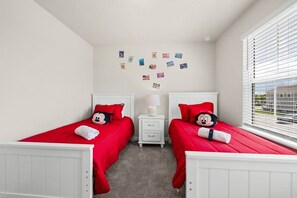Ein Schlafzimmer mit zwei Betten, jeweils mit roter Bettwäsche und Mickey Mouse Kissen, einem Nachttisch mit Lampe sowie einer Wand mit Fotos.