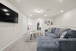 Ein modernes Wohnzimmer mit einem an der Wand befestigten Flachbildfernseher, einem grauen Sofa, einem Couchtisch und einem gemusterten Zierkissen.