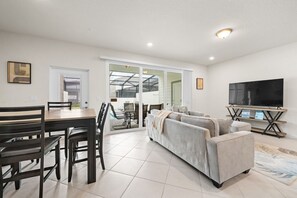 Ein modernes Wohnzimmer mit einer Couch, einem Esstisch mit Stühlen, einem Fernseher und einer Glastür, die auf eine Terrasse führt.