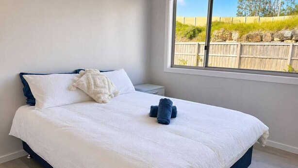 A neatly made bed with white bedding and a blue headboard in a room with a large window. - No.20 3B Pimpama Home (Pimpama)