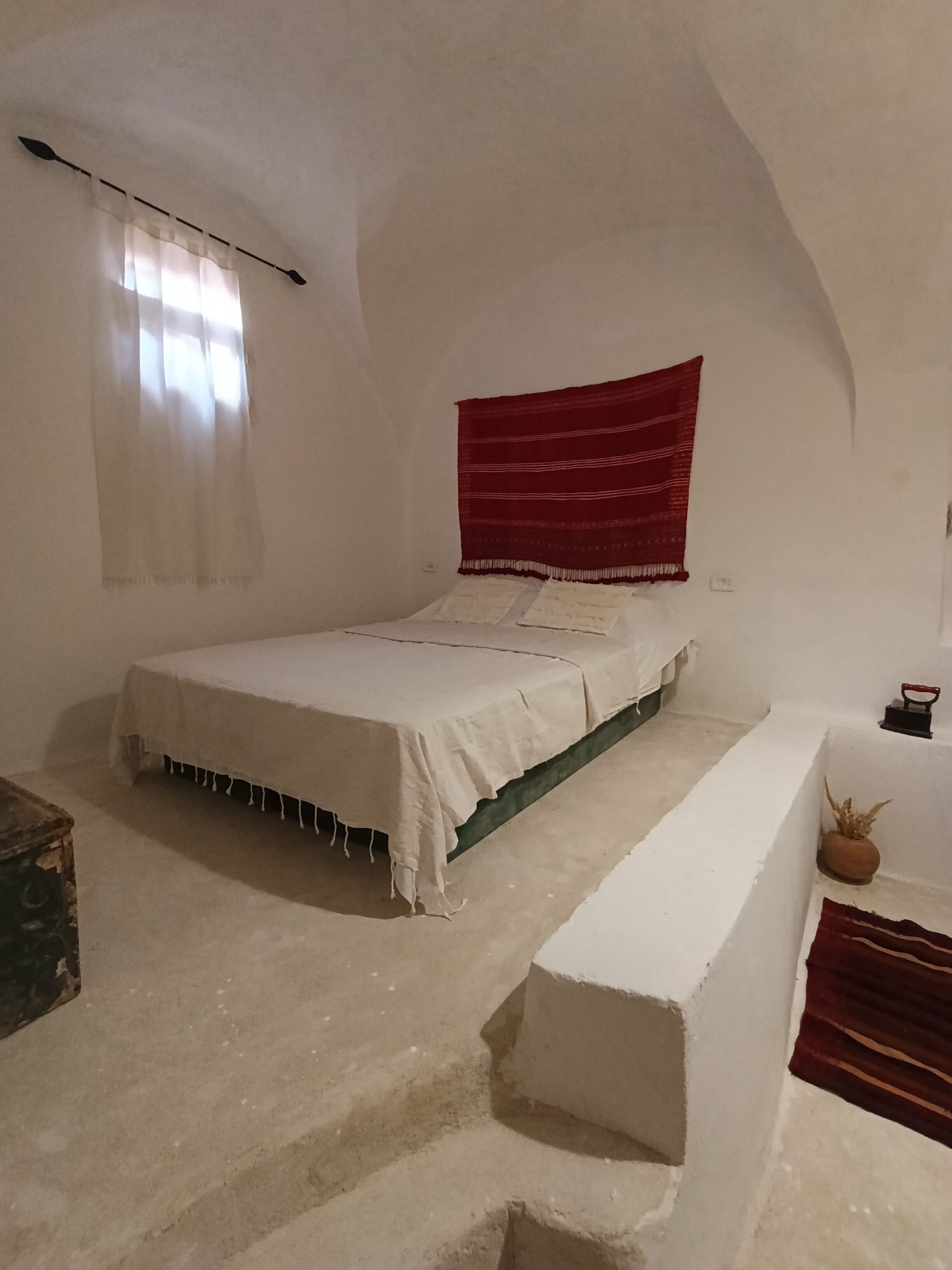 Ein Schlafzimmer mit einem Einzelbett, einer Bank, einem Fenster mit Vorhängen und einem roten Wandteppich.