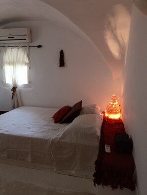 Un dormitorio con una cama, una mesita de noche, aire acondicionado de pared y una ventana con cortinas.