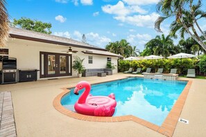 A swimming pool with a pink flamingo float, a barbecue area, and outdoor seating with umbrellas. - 3-bedroom house in Delray Beach with Jacuzzi, Cold Plunge, Sauna, Heated Pool (Delray Beach)