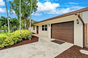 A single-story house with a brown garage door, a front porch, and a well-maintained lawn with yellow shrubs. - 3-bedroom house in Delray Beach with Jacuzzi, Cold Plunge, Sauna, Heated Pool (Delray Beach)